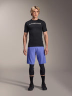 Alpinestars A-Aria Switch-tröja, kortärmad MTB-tröja, svart med spegelvänd vit Alpinestars-logotyp, bärs av en manlig modell med blå cykelshorts och svarta knäskydd, ventilerande funktionskläder för stigcykling