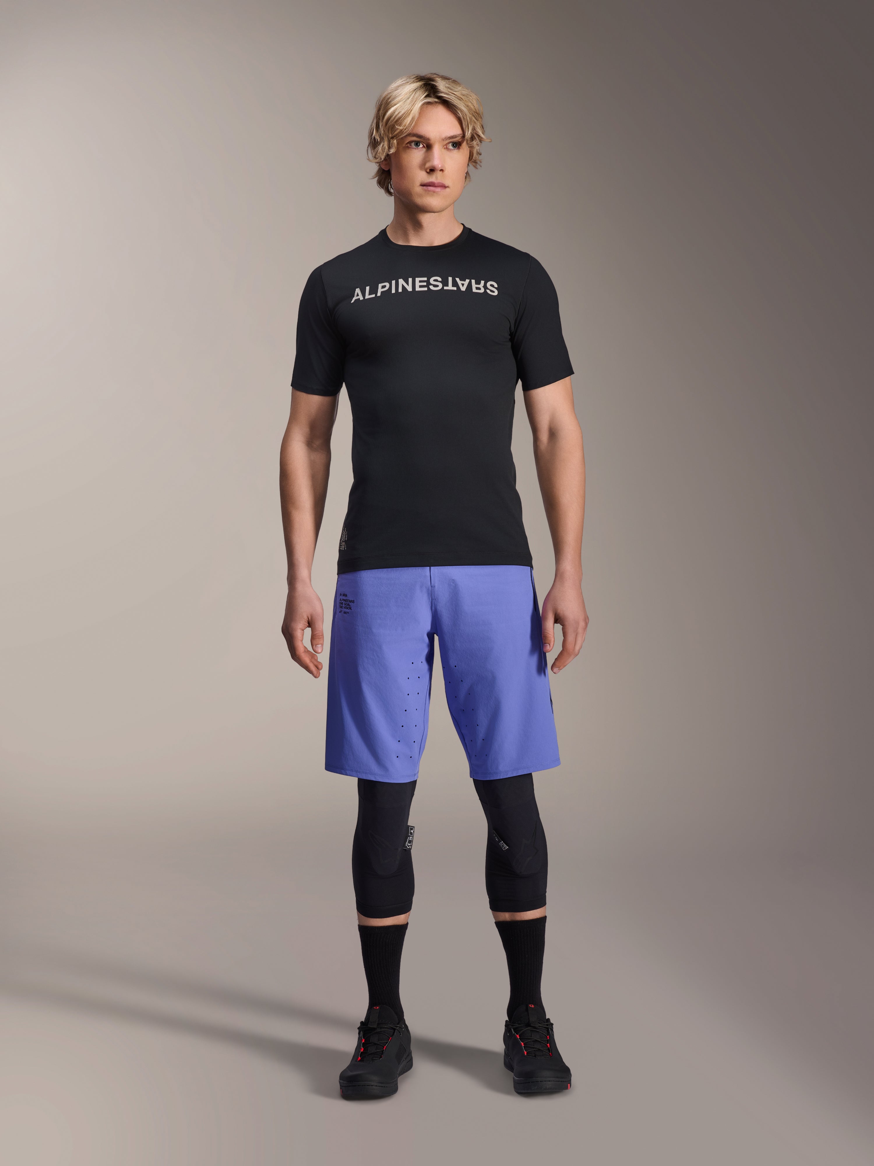 Alpinestars A-Aria Switch-tröja, kortärmad MTB-tröja, svart med spegelvänd vit Alpinestars-logotyp, bärs av en manlig modell med blå cykelshorts och svarta knäskydd, ventilerande funktionskläder för stigcykling