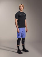 Manlig modell bär Alpinestars mountainbike-kläder, med den svarta A-Aria Switch SS-tröjan och Baja Blue A-Aria Lite-shorts, kombinerat med svarta knäskydd, svarta strumpor och svarta mountainbike-skor i en studiobild i helfigur.