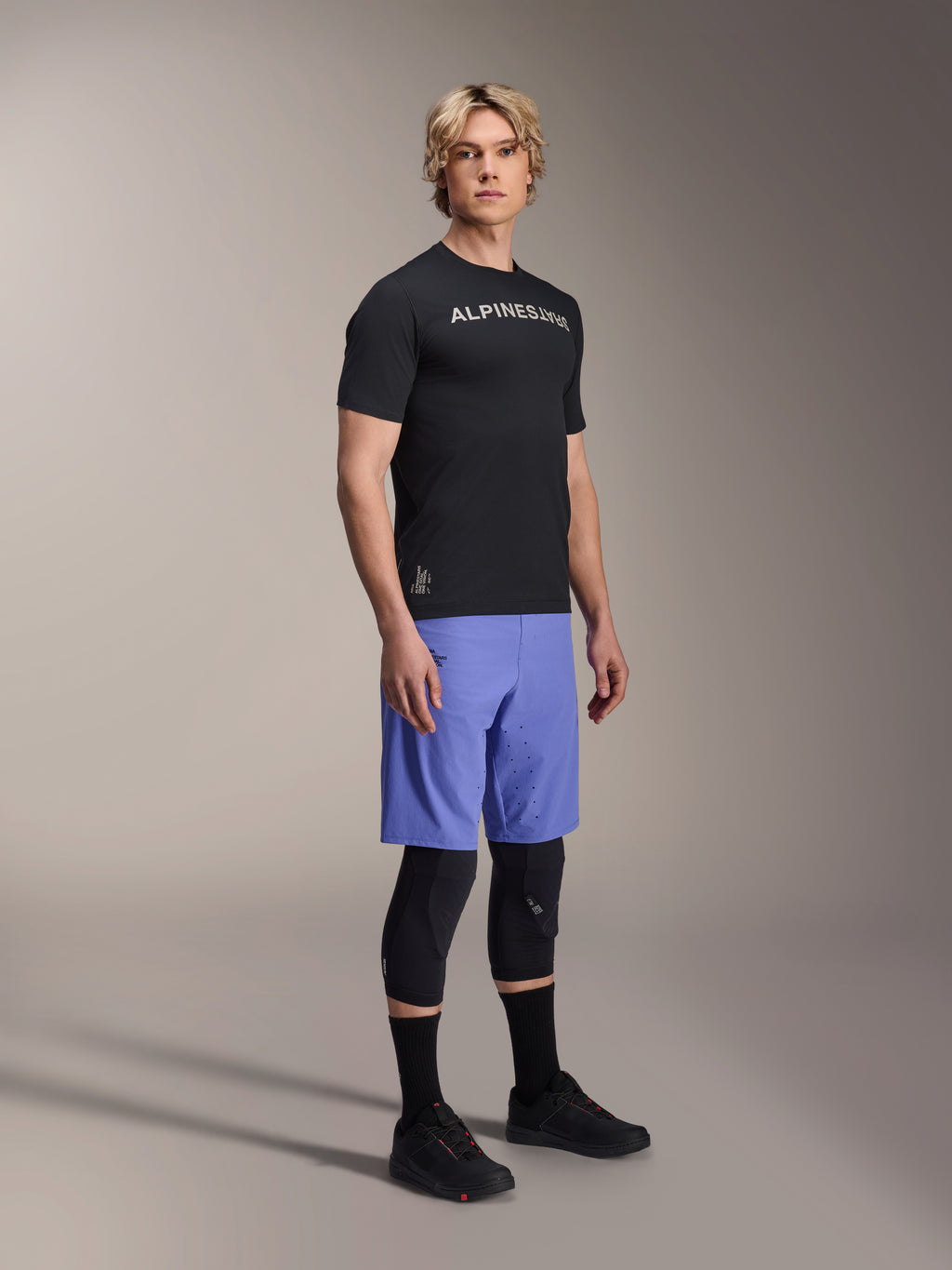 Manlig modell i Alpinestars A-Aria Switch kortärmad tröja i svart med vit logotyp på bröstet, ljusblå mountainbike-shorts, svarta knäskydd och svarta cykelskor, helkroppsbild i studio för MTB-utrustning