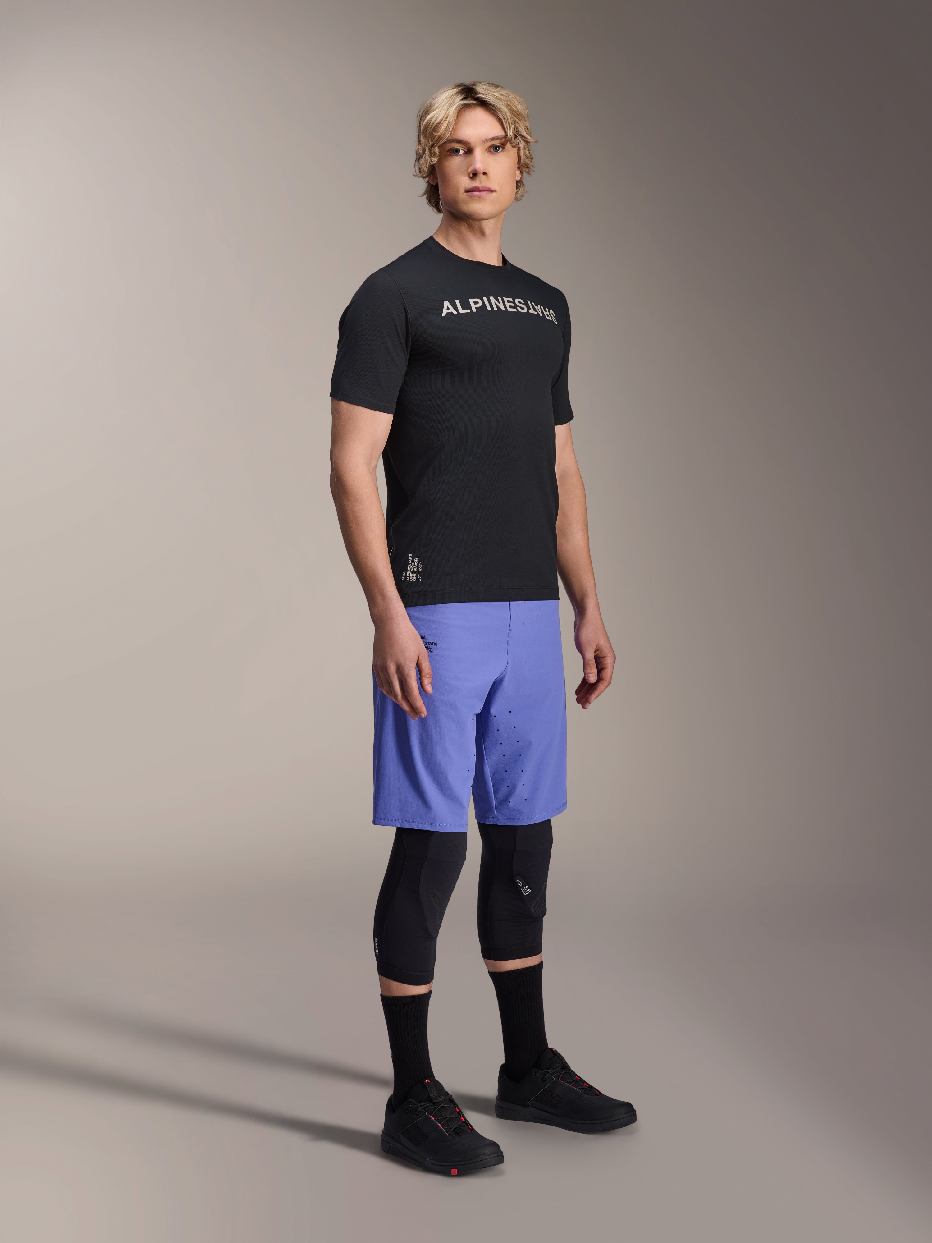 Manlig modell i Alpinestars A-Aria Switch kortärmad tröja i svart med vit logotyp på bröstet, ljusblå mountainbike-shorts, svarta knäskydd och svarta cykelskor, helkroppsbild i studio för MTB-utrustning