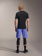 Alpinestars A-Aria Switch kortärmad tröja, svart MTB-tröja, sedd bakifrån, kombinerad med blå mountainbike-shorts och svarta benskydd, teknisk cykelutrustning för trail-cykling
