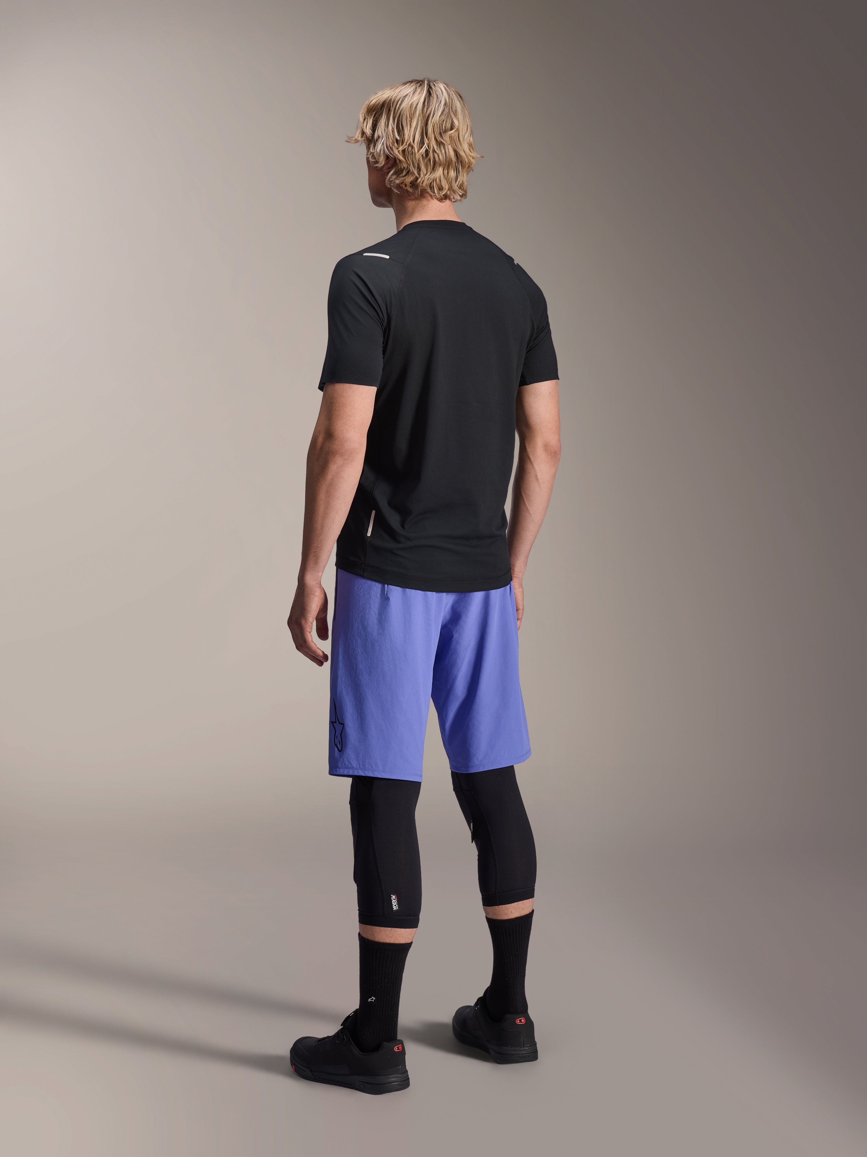 Alpinestars A-Aria Switch Jersey, kortärmad MTB-tröja, svart, baksida på modell med blå shorts och svarta knäskydd, teknisk cykelutrustning för stigcykling
