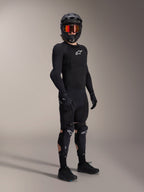 Alpinestars A-Impact Plasma Pro Knee Shin Protectors, Black and White, worn by a rider with a complete mountain bike protective outfit, including a black long-sleeve armored top, black gloves, and a full-face helmet with orange goggles.