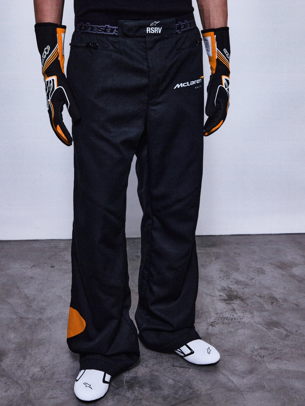 Alpinestars RSRV Arena Suit Pants, svarta racingbyxor med McLaren-logotyp, orangea detaljer på sidan och knä, person iklädd med svarta och orangea handskar samt vita racingskor