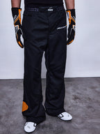 Alpinestars RSRV Arena Suit Pants, svarta racingbyxor med McLaren-logotyp, orangea detaljer på sidan och knä, person iklädd med svarta och orangea handskar samt vita racingskor
