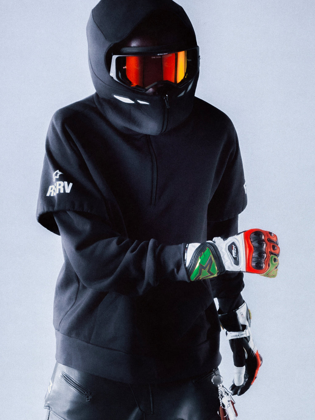 Alpinestars RSRV Vault Hoodie, svart premiumhuvtröja med huva, unisex streetwear, vit logotyp på ärmen, visad med motorcykelhjälm och reflekterande orange visir, färgglada handskar, modern skyddsutrustning