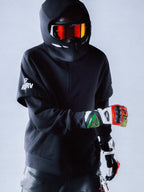 Alpinestars RSRV Vault Hoodie, svart premiumhuvtröja med huva, unisex streetwear, vit logotyp på ärmen, visad med motorcykelhjälm och reflekterande orange visir, färgglada handskar, modern skyddsutrustning