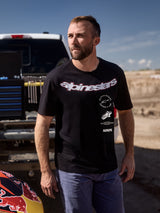 Alpinestars Abus CSF Tee, Short Sleeve T-Shirt, Black, featuring a large curved Alpinestars wordmark and multi-stacked logos, worn by a man in a motorsport paddock setting, casual lifestyle streetwear