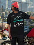 Alpinestars Aplomb Oversized CSF Tee, kortärmad t-shirt, svart, med en stor turkos och vit färggradient Astars-logotyp och målflaggsgrafik, bärs med en korallfärgad mössa, stående bredvid en röd motorcykel, avslappnad lifestyle streetwear