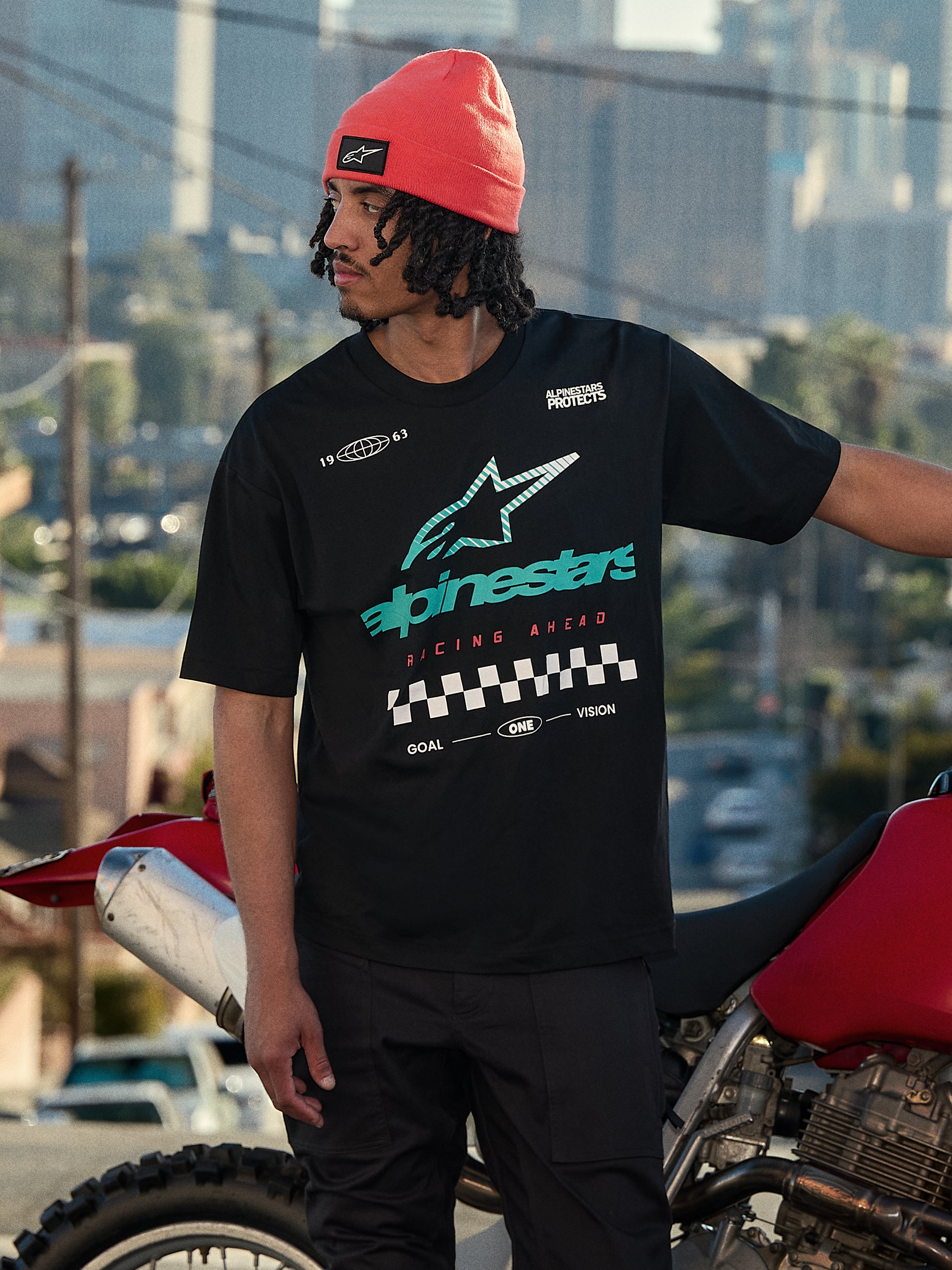 Alpinestars Aplomb Oversized CSF Tee, kortärmad t-shirt, svart, med en stor turkos och vit färggradient Astars-logotyp och målflaggsgrafik, bärs med en korallfärgad mössa, stående bredvid en röd motorcykel, avslappnad lifestyle streetwear