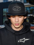 Alpinestars Contender Trucker Hat, svart lifestyle-keps med mesh-baksida, strukturerad krona med ett broderat Astars-logopatch, bärs av en man i en svart Alpinestars-hoodie, avslappnad motorsport-inspirerad streetwear.