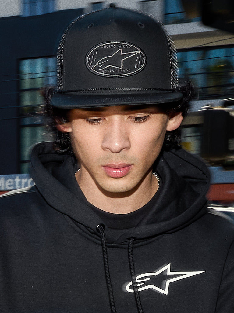 Alpinestars Contender Trucker Hat, black lifestyle cap with poly mesh back, structured crown featuring an embroidered Astars logo patch, worn by a man with a black Alpinestars hoodie, casual motorsport-inspired streetwear.