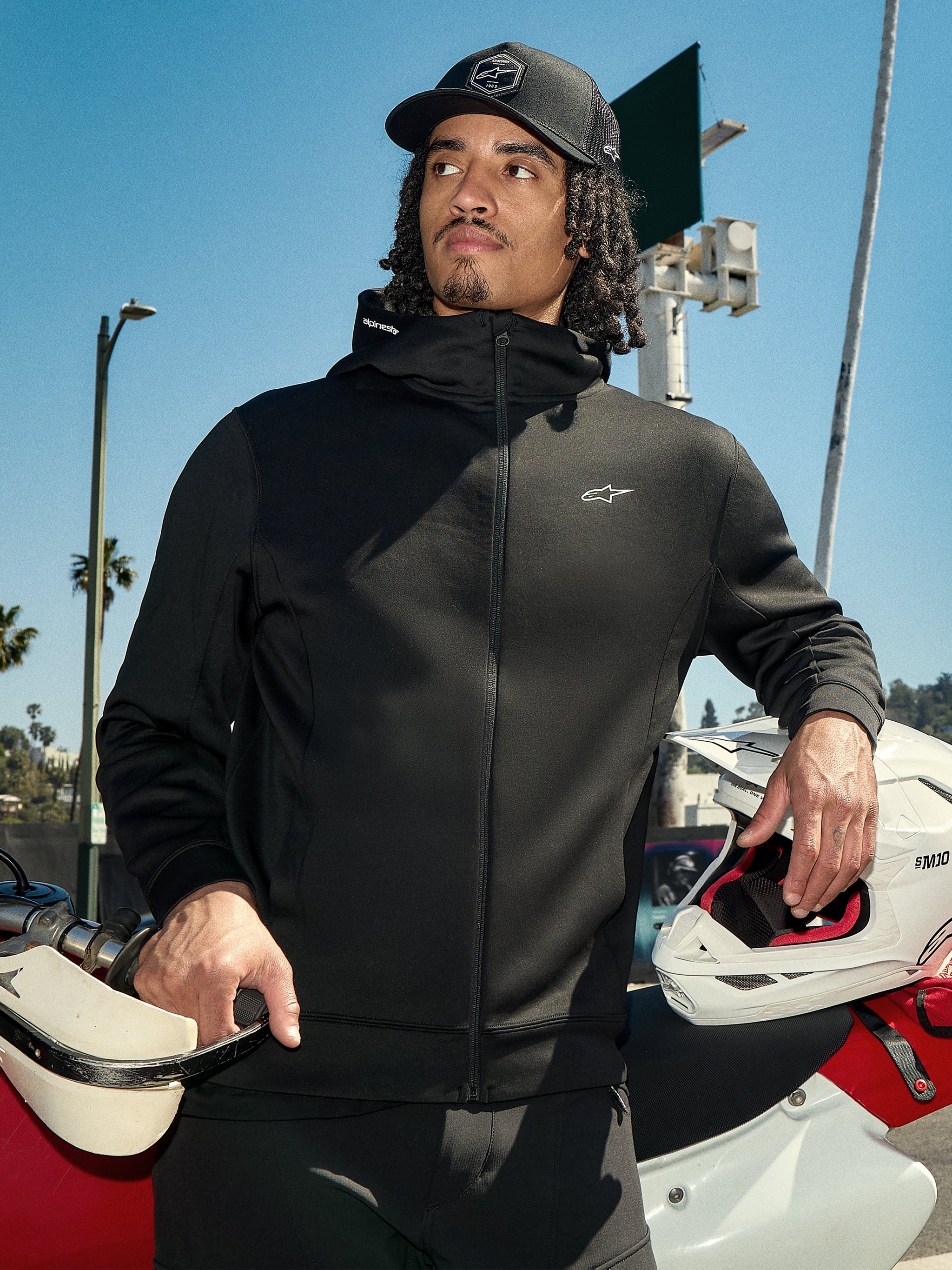 Alpinestars Cypher Hoodie, svart fleecehuvtröja med dragkedja, hög huva med logotyp, liten vit Astars-logotyp på bröstet, bärs med en svart keps av en förare som håller en vit motocrosshjälm, avslappnad motorsport-streetwear