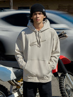 Alpinestars Emergent Oversized Hoodie i Off White, oversized fleece-huvtröja med 3D-broderad Astars-logotyp, känguruficka och huva med dragsko, bärs av en man i en svart mössa bredvid en motorcykel, casual lifestyle motorsport streetwear