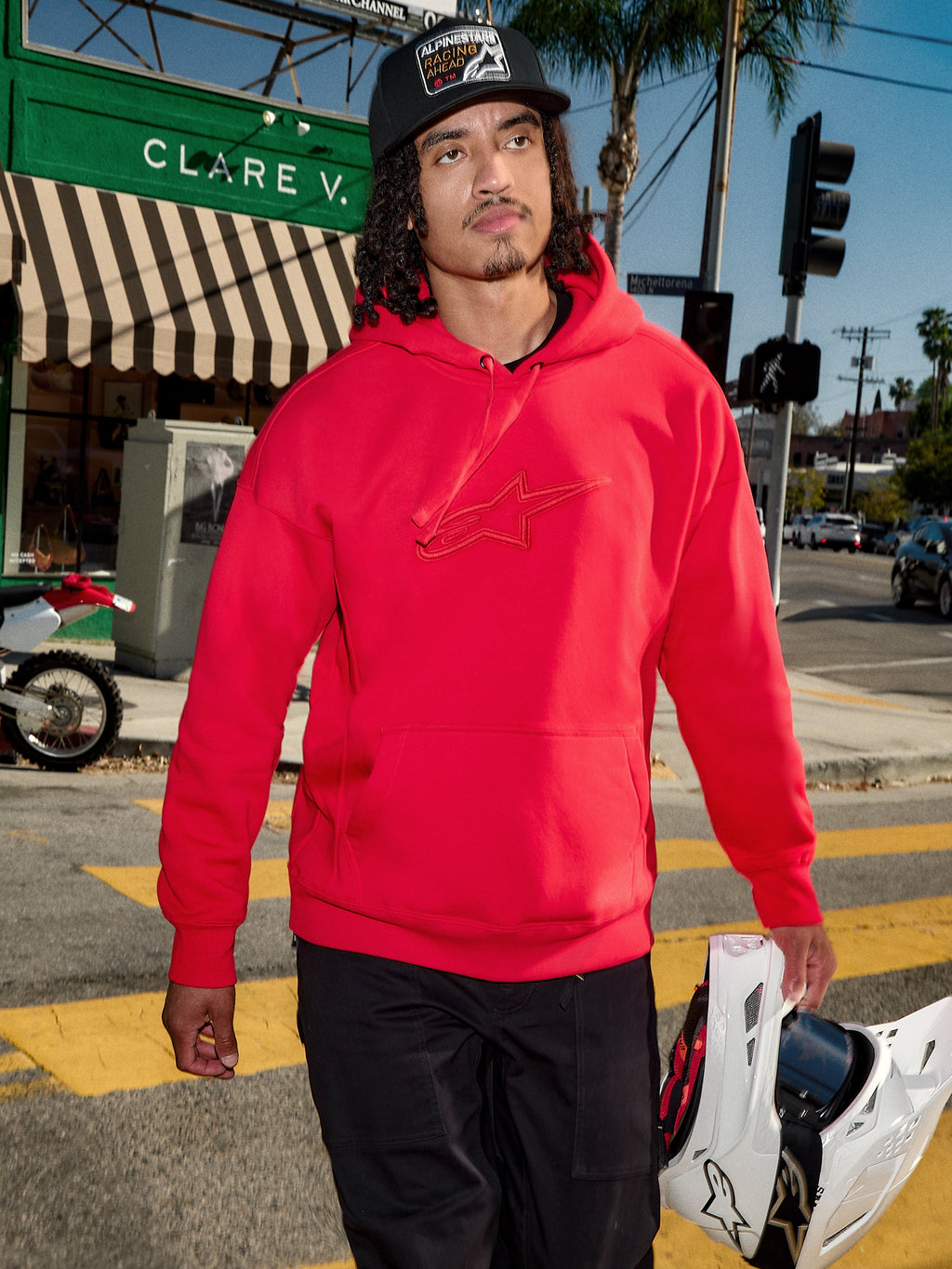 Alpinestars Emergent Oversized Hoodie, röd, 3D-broderad Astars-logotyp på bröstet, bärs med svarta byxor och en baseballkeps tillsammans med en vit motocrosshjälm, avslappnad urban streetwear-stil