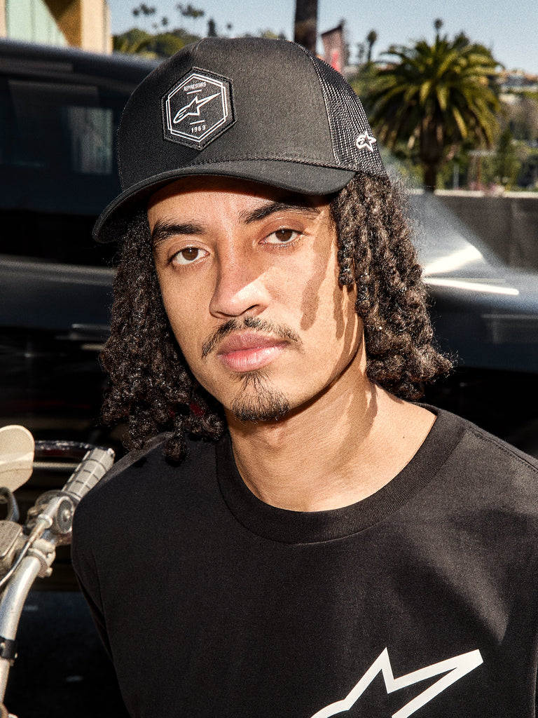 Alpinestars Hexeon Trucker Hat, svart truckerkeps, livsstilsbild av en man som bär kepsen med böjd skärm, hexagonal Astars-logopatch och mesh-baksida, matchad med en svart t-shirt med logotyp, avslappnad motorsport-inspirerad streetwear