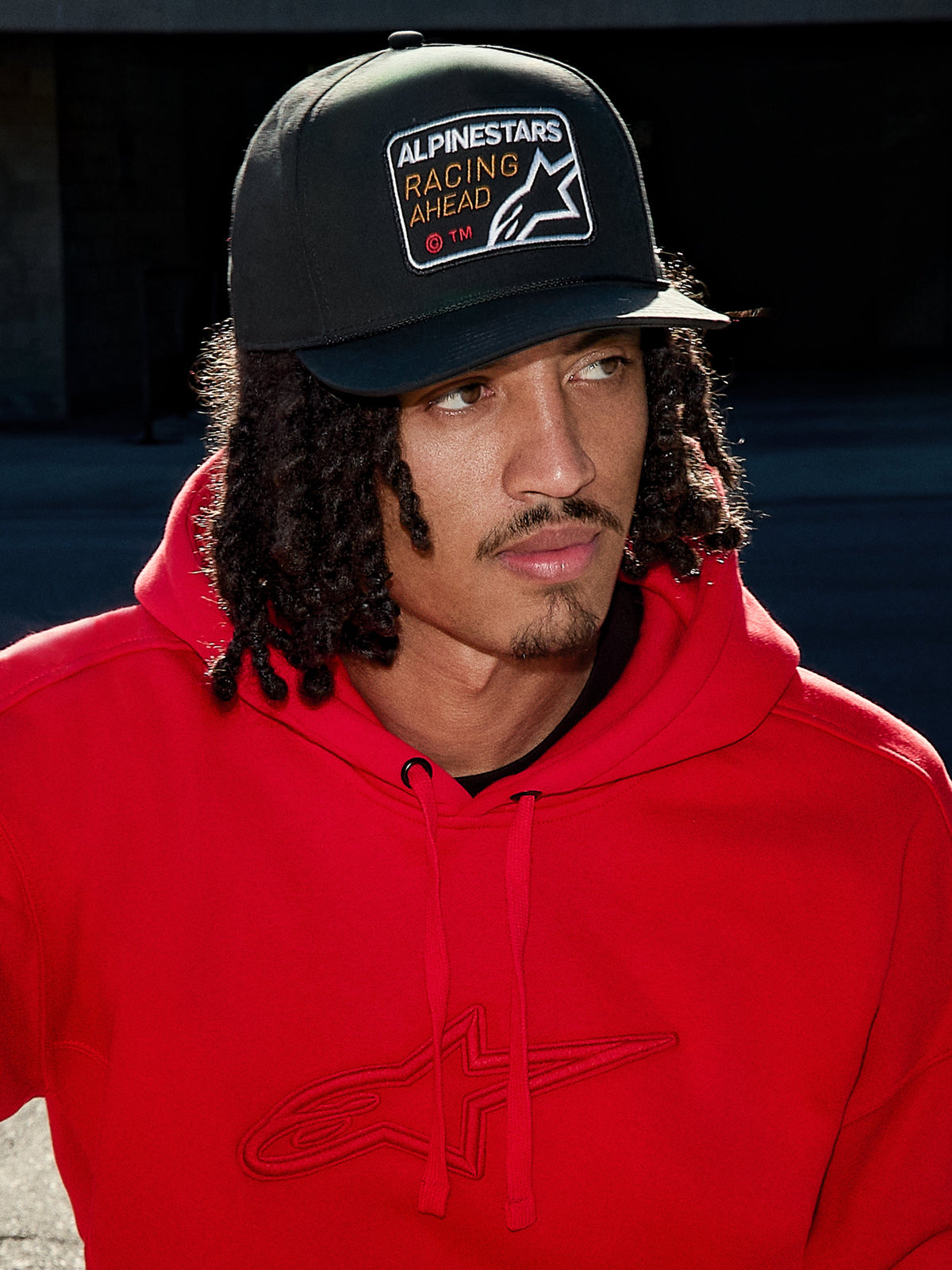 Man som bär Alpinestars Intuitive Snapback-keps i svart, med strukturerad krona och broderat 'Racing Ahead'-logopatch, kombinerat med en klarröd hoodie, avslappnad lifestyle-streetwear