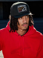 Man som bär Alpinestars Intuitive Snapback-keps i svart, med strukturerad krona och broderat 'Racing Ahead'-logopatch, kombinerat med en klarröd hoodie, avslappnad lifestyle-streetwear