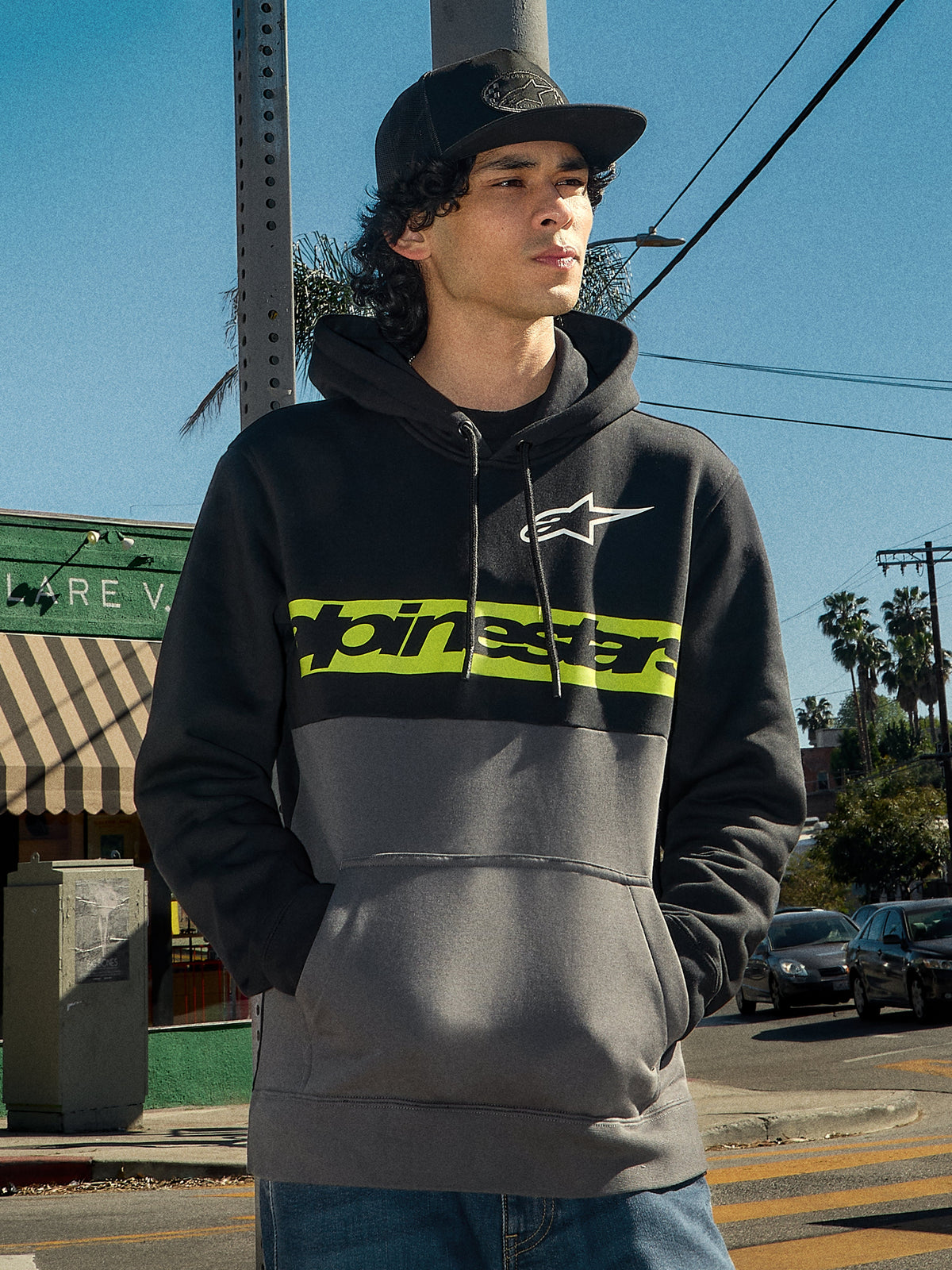 Alpinestars Parallel Hoodie, lifestyle streetwear, färgblockerad design i svart och gunmetal, limegrönt bröstband med Alpinestars logotyp, vit Astars-logotyp, huva med dragsko, bärs av en man i en urban miljö