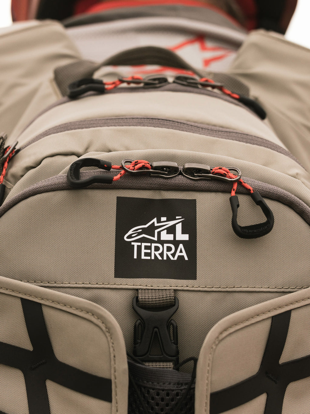 Alpinestars AT-15 ryggsäck, Aluminum Walnut, närbild på All Terra-logotypmärke på beige teknisk textil, med orangea snörade dragkläppar och robusta konstruktionsdetaljer