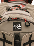 Alpinestars AT-15 ryggsäck, Aluminum Walnut, närbild på All Terra-logotypmärke på beige teknisk textil, med orangea snörade dragkläppar och robusta konstruktionsdetaljer