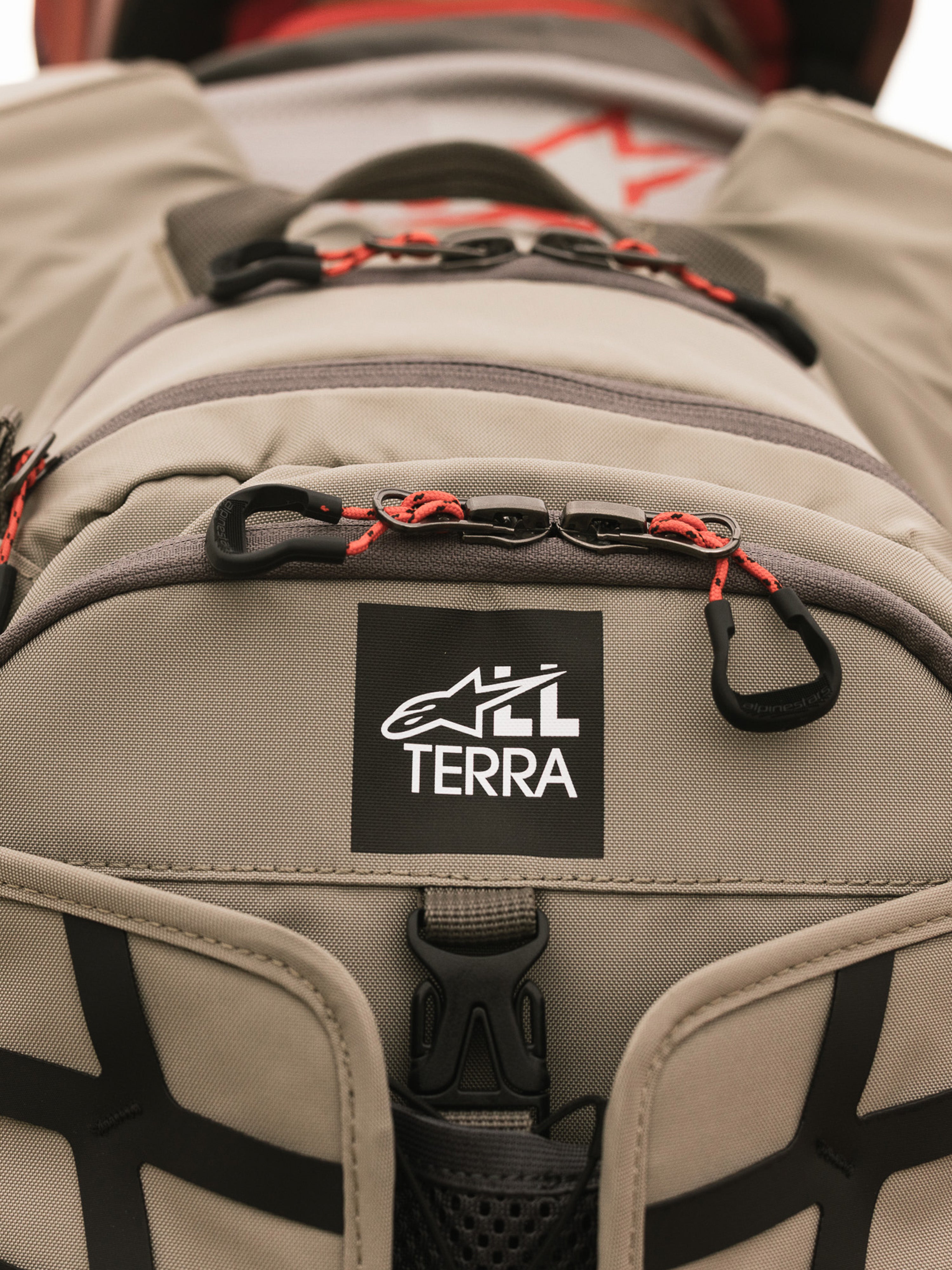 Alpinestars AT-15 ryggsäck, Aluminum Walnut, närbild på All Terra-logotypmärke på beige teknisk textil, med orangea snörade dragkläppar och robusta konstruktionsdetaljer