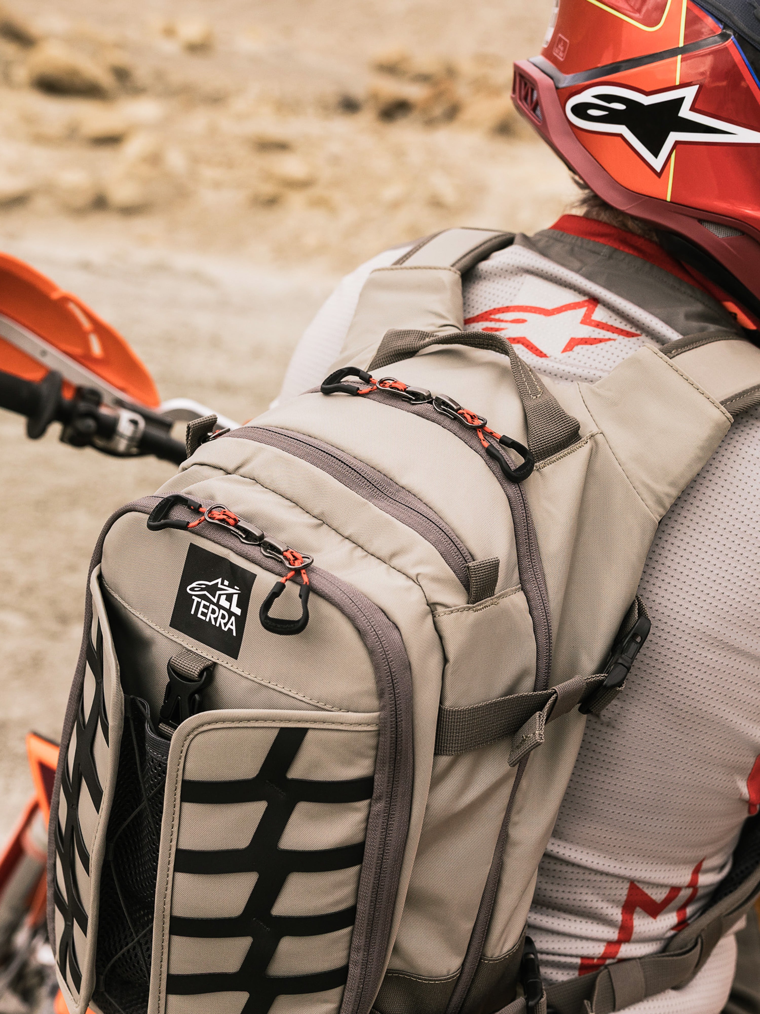 Alpinestars AT-15 Back Pack, äventyrsryggsäck för motorcykel, Aluminum Walnut och svart, buren av en förare i en ökenmiljö, med röda dragkläppar, TERRA-logotyp och svarta externa remmar, matchad med en röd hjälm och vit Alpinestars-tröja