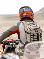 Alpinestars AT-15 ryggsäck i Aluminum Walnut, bärs av en äventyrsmotorcyklist i ett ökenlandskap, robust beige textilkonstruktion med svarta webbing-detaljer, orange dragkedjor och 'All Terra'-logotyp, kombinerad med röd och orange hjälm och grå MC-utrustning för terrängkörning