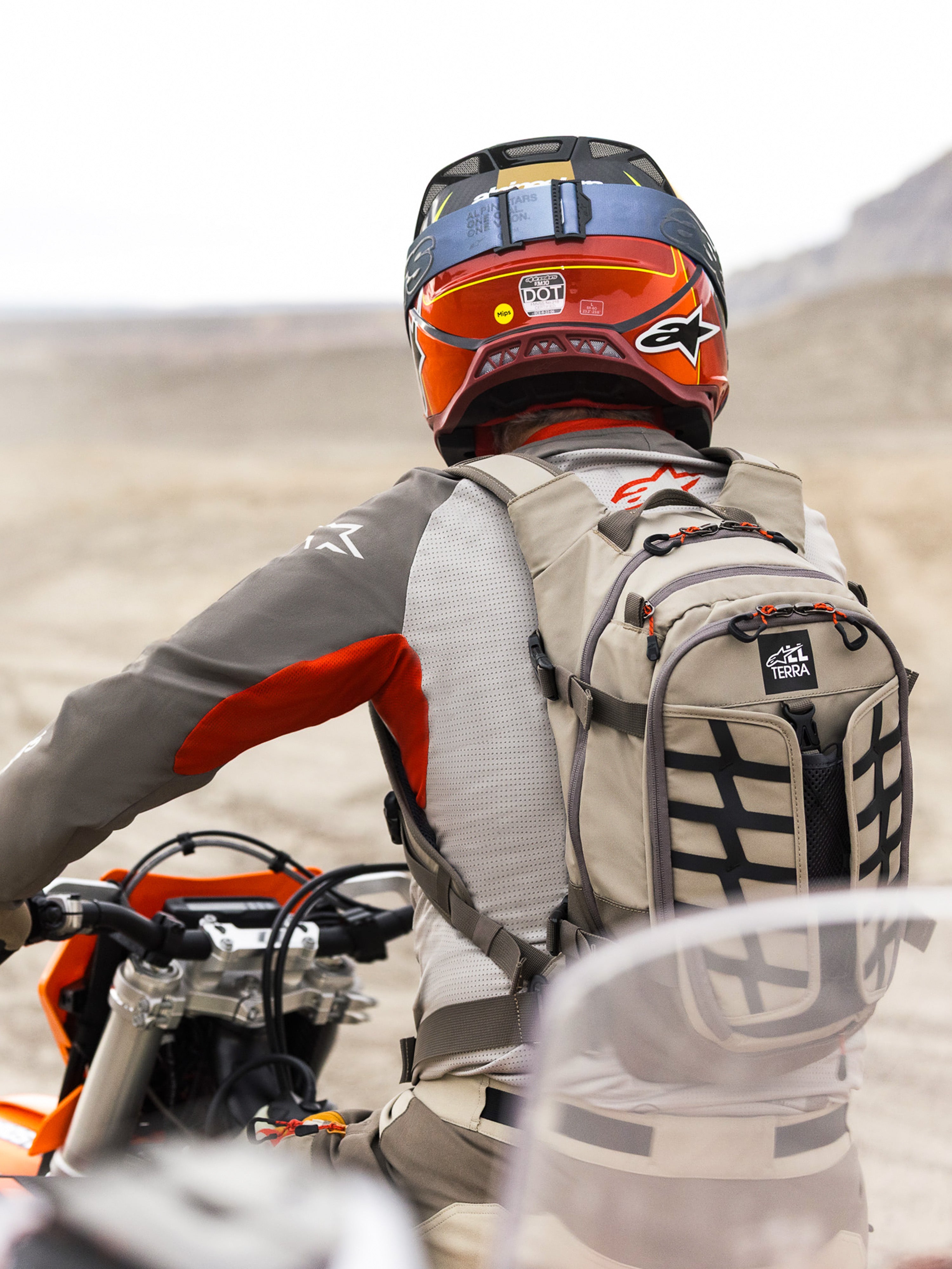 Alpinestars AT-15 ryggsäck i Aluminum Walnut, bärs av en äventyrsmotorcyklist i ett ökenlandskap, robust beige textilkonstruktion med svarta webbing-detaljer, orange dragkedjor och 'All Terra'-logotyp, kombinerad med röd och orange hjälm och grå MC-utrustning för terrängkörning