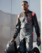 Gokartförare som bär Alpinestars KMX-9 V3 Graphic 6 Suit i Anthracite Black och rött, håller en mattsvart hjälm och handskar, med tydliga röda Alpinestars-logotyper och en stänkmönstrad grafisk design, utomhus motorsportmiljö.