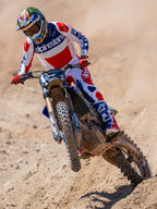 Motocross rider in action wearing Alpinestars Limited Edition Fortitude Tech 10 Boots, Bright Red White and Blue racing jersey and pants, coordinated patriotic gear set for off-road performance on a dirt track.