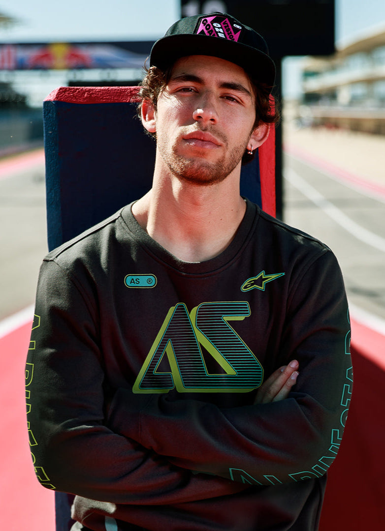 Alpinestars Ayess Csf Tee, långärmad t-shirt, svart, med grön och gul tonad AS-grafik på bröstet och ärmarna, avslappnat motorsportinspirerat streetwear buren på en racerbana