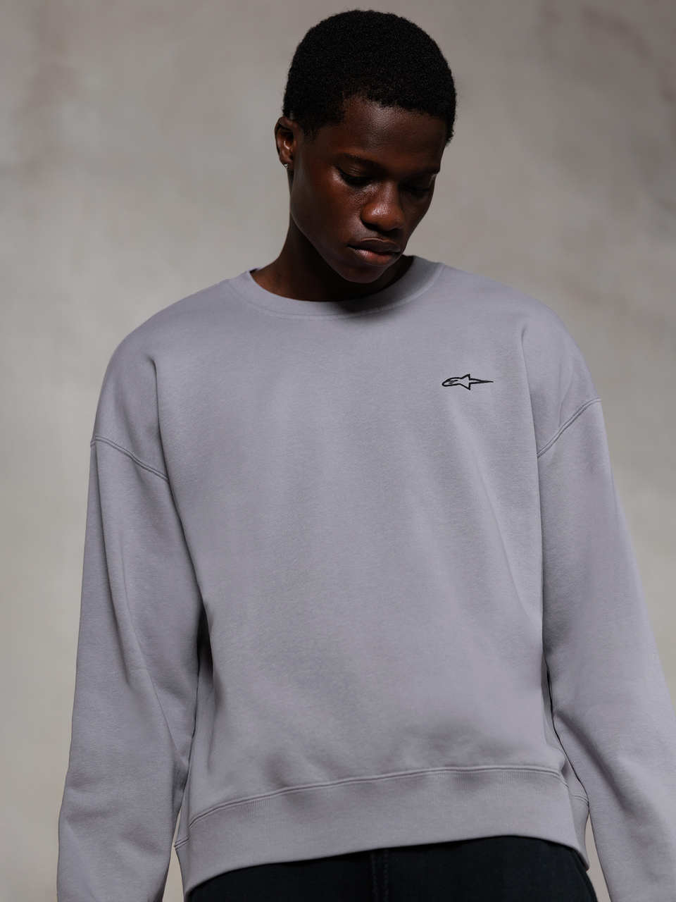 Alpinestars Essentials Ageless Crew, silverfärgad premium crewneck-tröja, med en liten svart Alpinestars-logotyp på bröstet, bärs av en modell, avslappnad lifestyle-streetwear med en mjuk fleecestruktur.