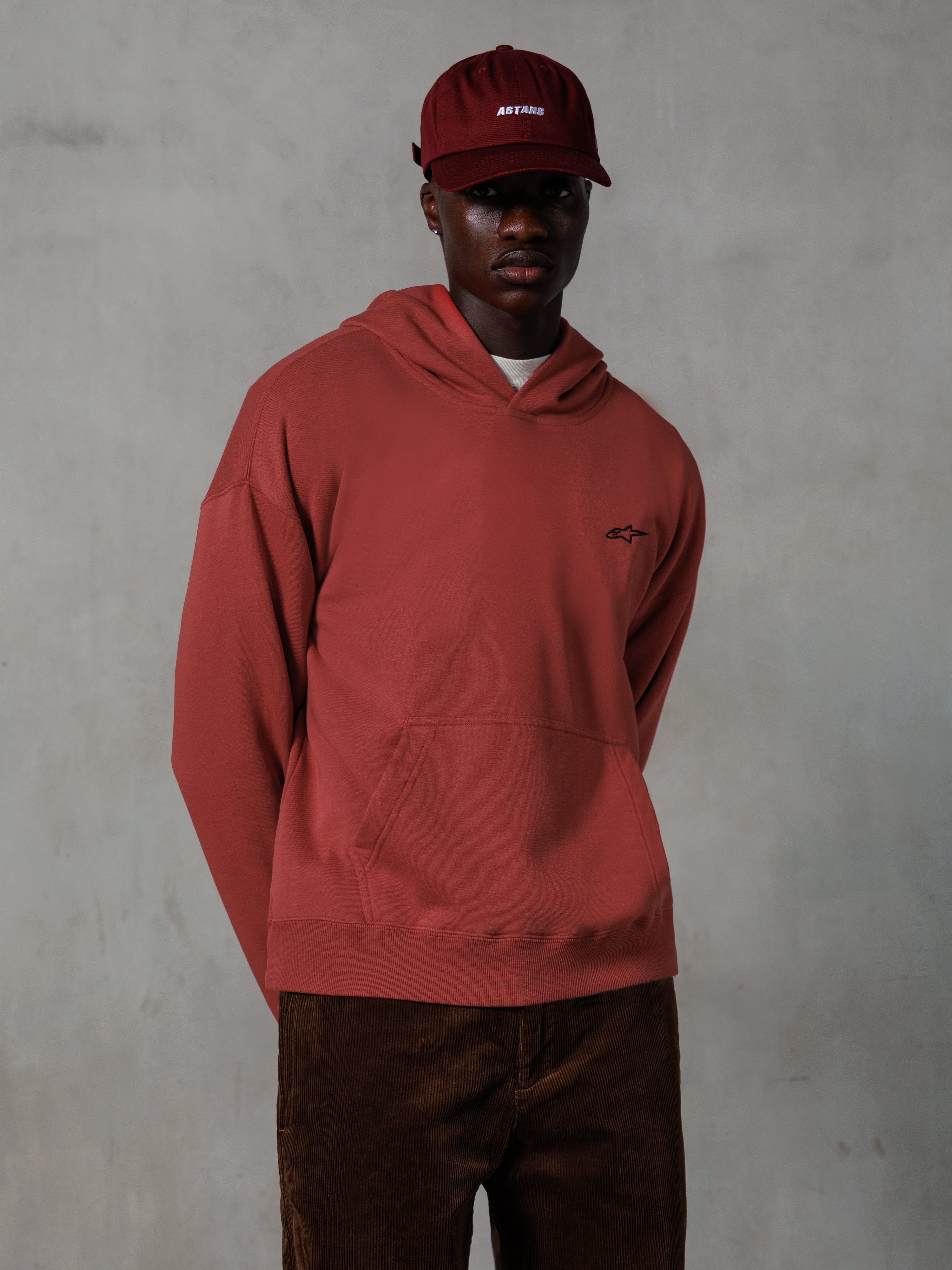 Alpinestars Essentials Ageless Hoodie, casual streetwear, Brick red, small black chest logo, front kangaroo pocket, worn with a matching red ASTARS cap and brown corduroy pants, clean minimalist design