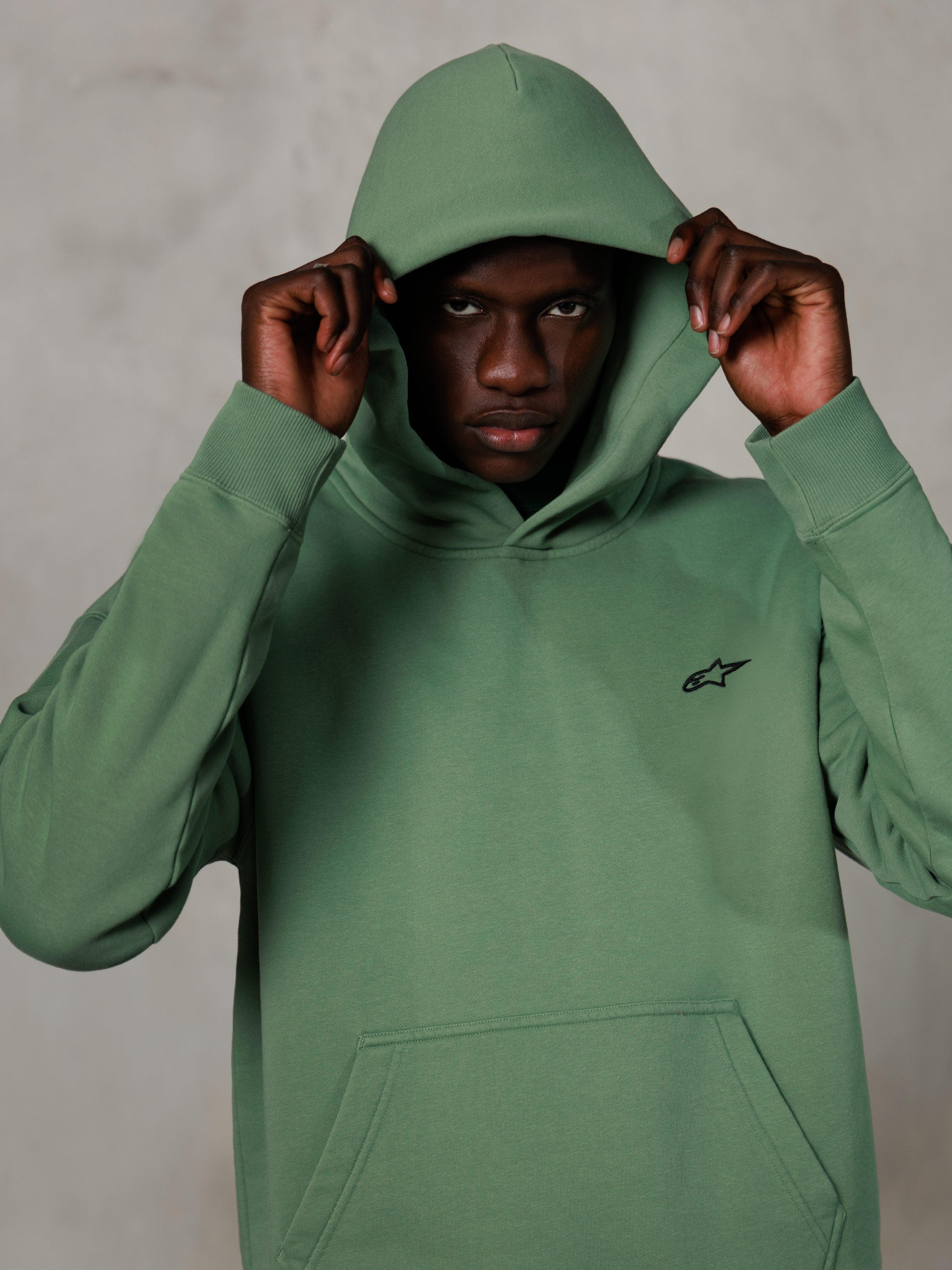 Alpinestars Essentials Ageless Hoodie, casual streetwear, kaktusgrön med en liten svart logotyp på bröstet, modell bär huvtröjan med huvan uppfälld, känguruficka fram, avslappnad motorsportinspirerad design