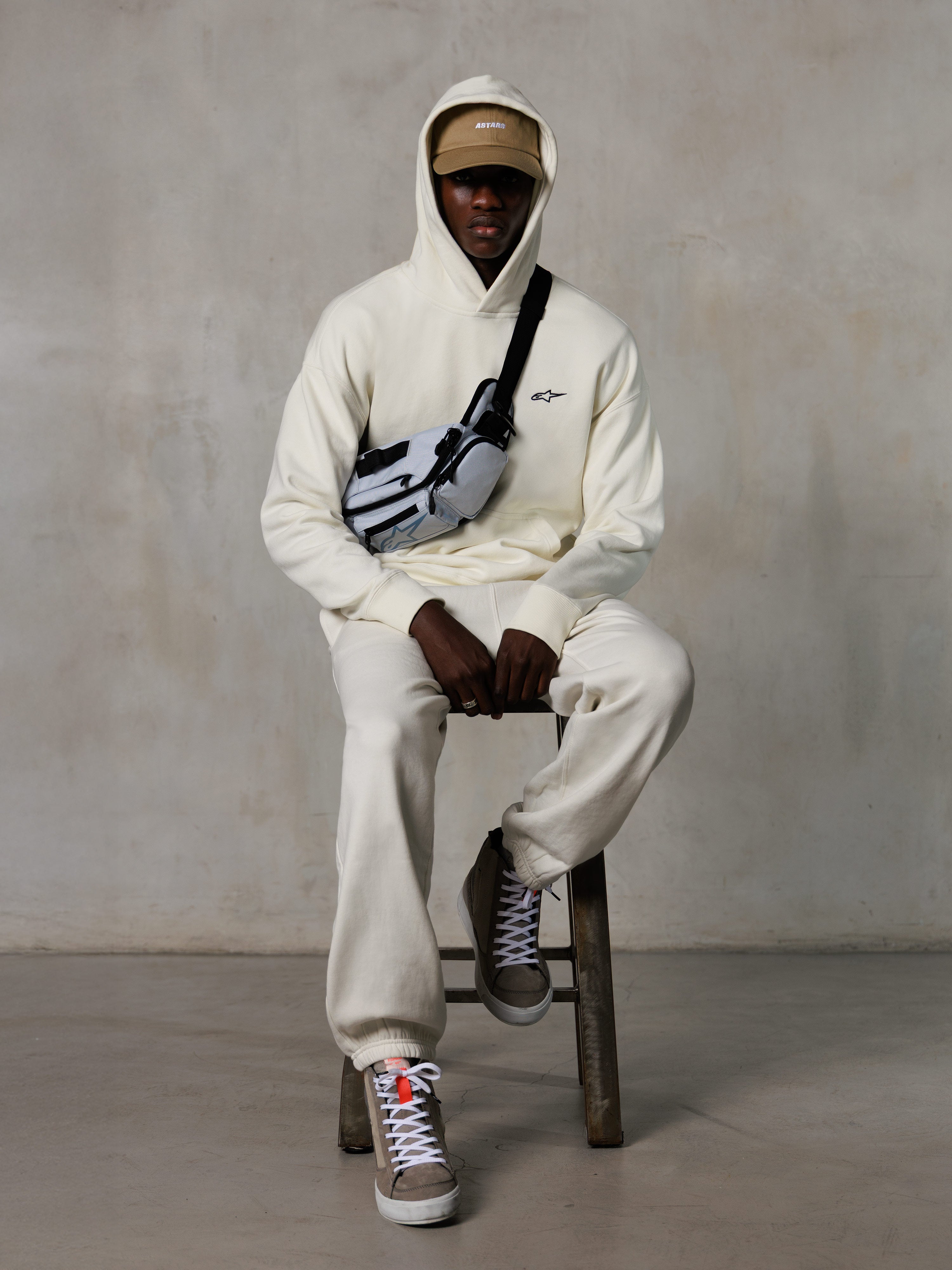 Alpinestars Essentials Ageless Hoodie, Off-White, worn by a model sitting on a stool with matching fleece joggers, tan ASTARS cap, and cross-body bag, casual motorsport-inspired streetwear