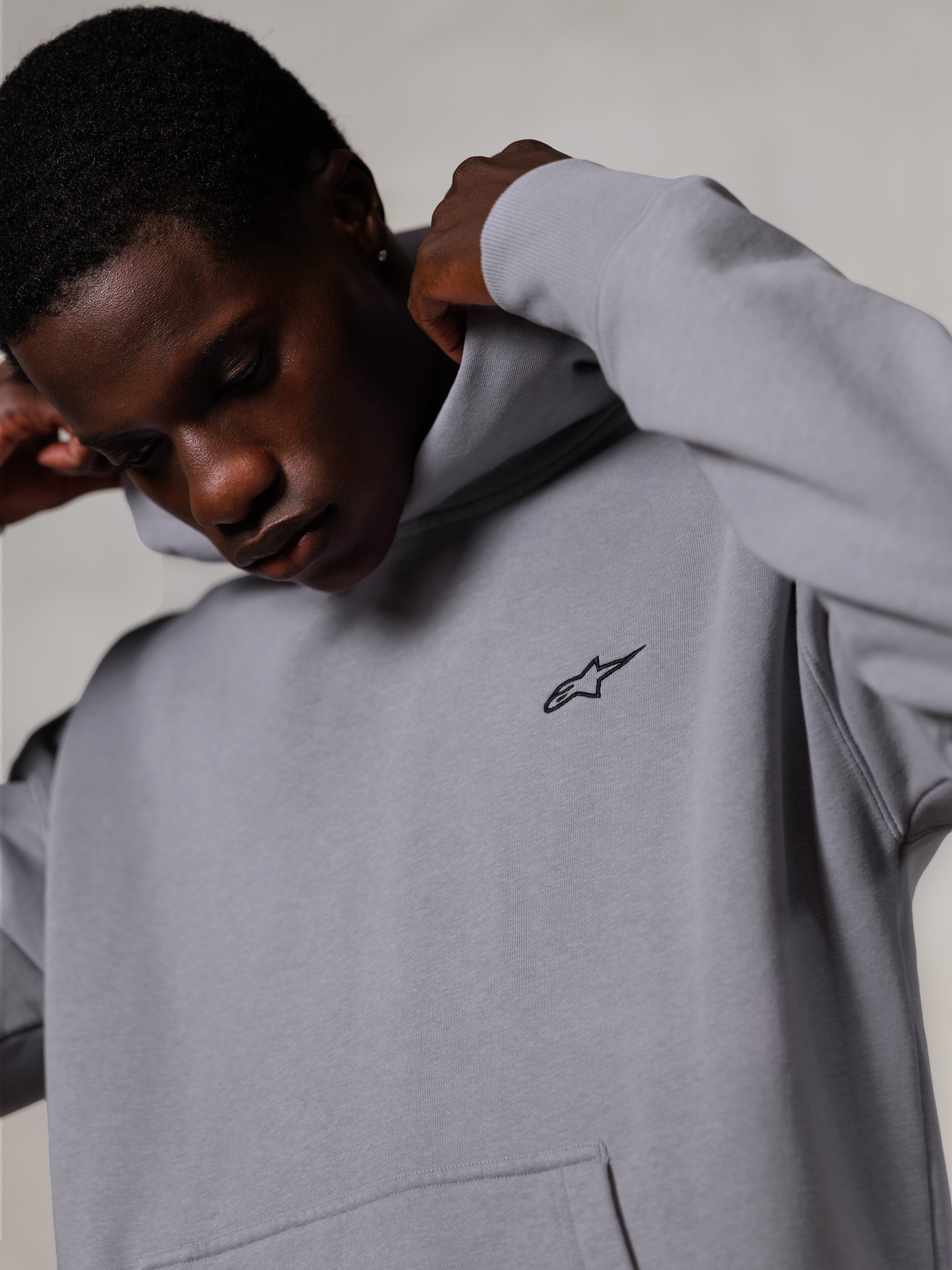 Alpinestars Essentials Ageless Hoodie, Silver, närbild på en man som justerar huvan på den grå huvtröjan, liten svart Alpinestars-logotyp på bröstet, premium motorsportinspirerade vardagskläder
