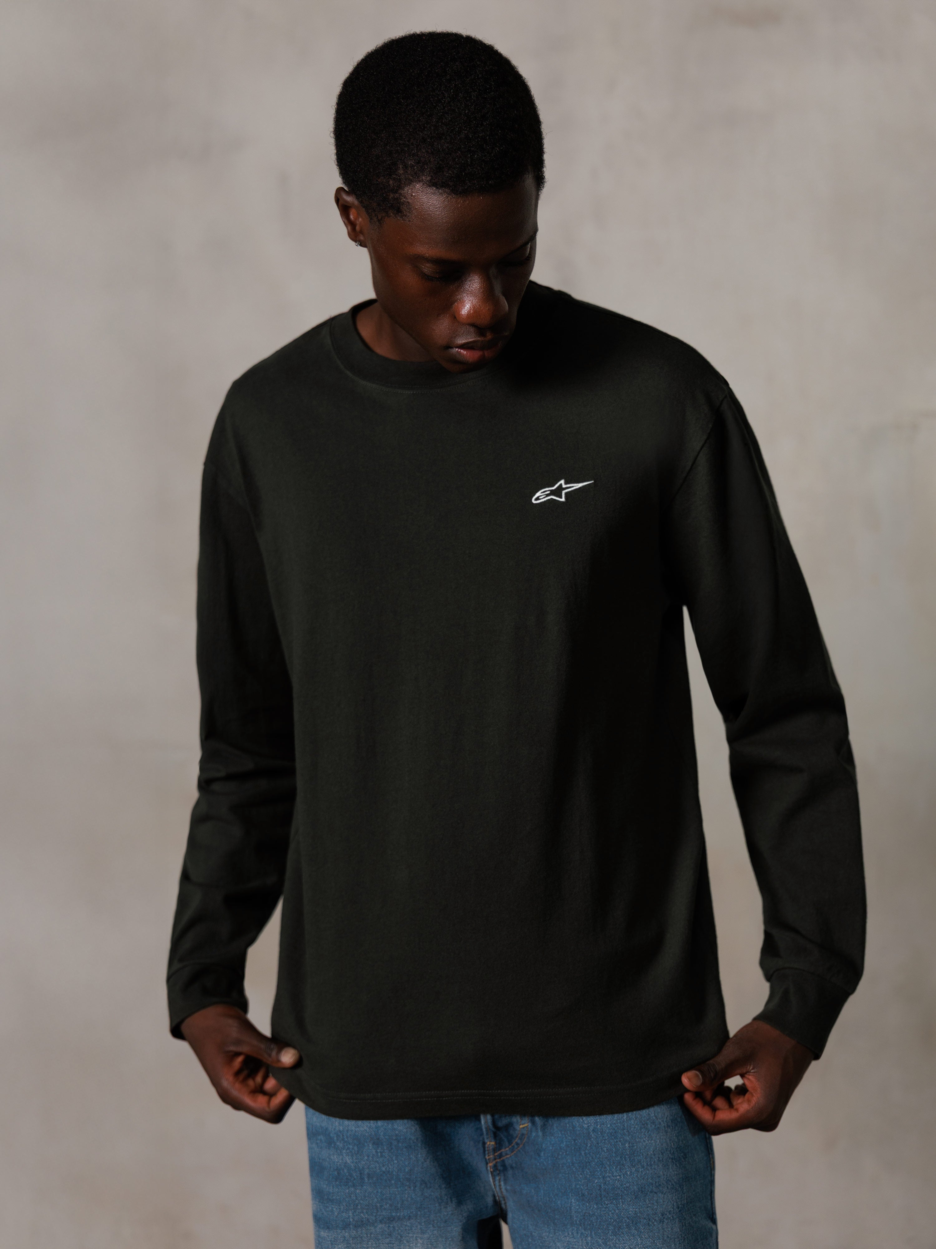 Alpinestars Essentials Ageless LS Tee, långärmad t-shirt, Off Black, med en liten vit logotyp på bröstet, bärs av en modell med blå jeans, avslappnad lifestyle-streetwear
