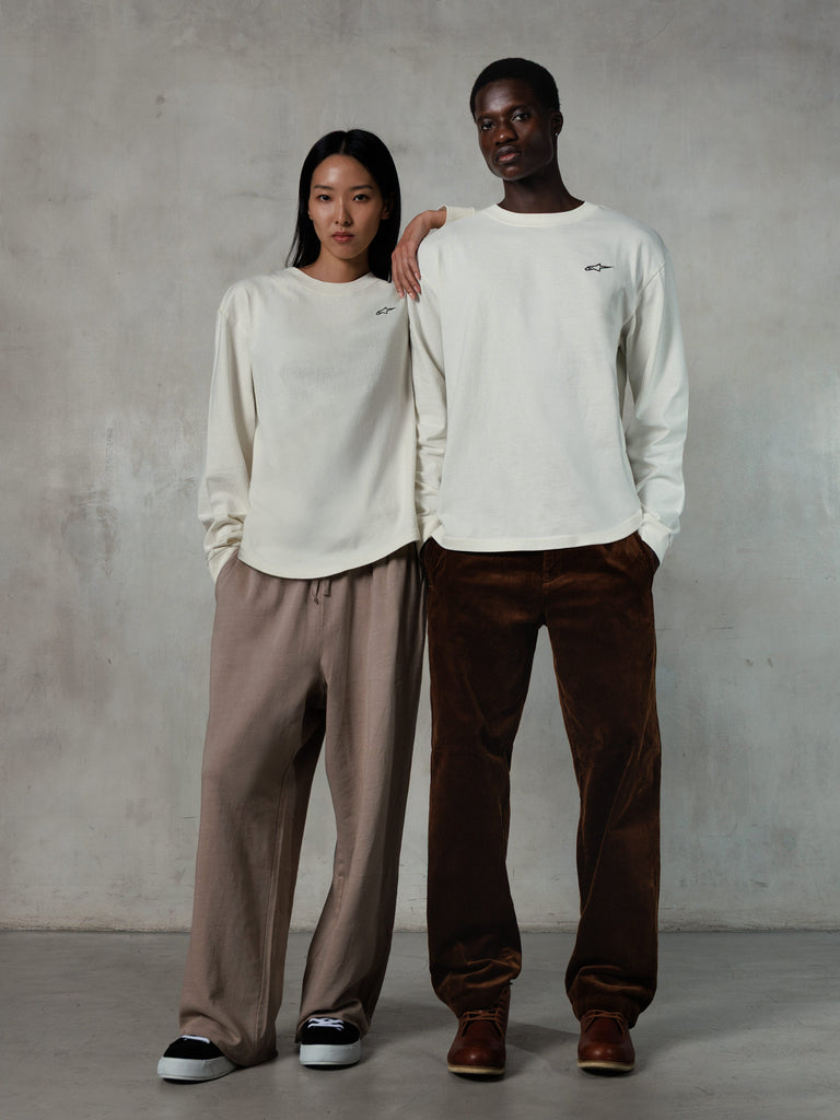 Alpinestars Essentials Ageless LS Tee, Long-Sleeve T-Shirt, Off White, worn by male and female models, small black Alpinestars logo on the chest, relaxed fit, styled with tan sweatpants and brown corduroy pants, casual motorsport-inspired streetwear