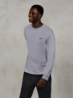Alpinestars Essentials Ageless LS Tee, långärmad t-shirt, silver, liten svart logotyp på bröstet, bärs av en leende modell med mörka byxor, avslappnad modern passform, vardagliga motorsport-livsstilskläder