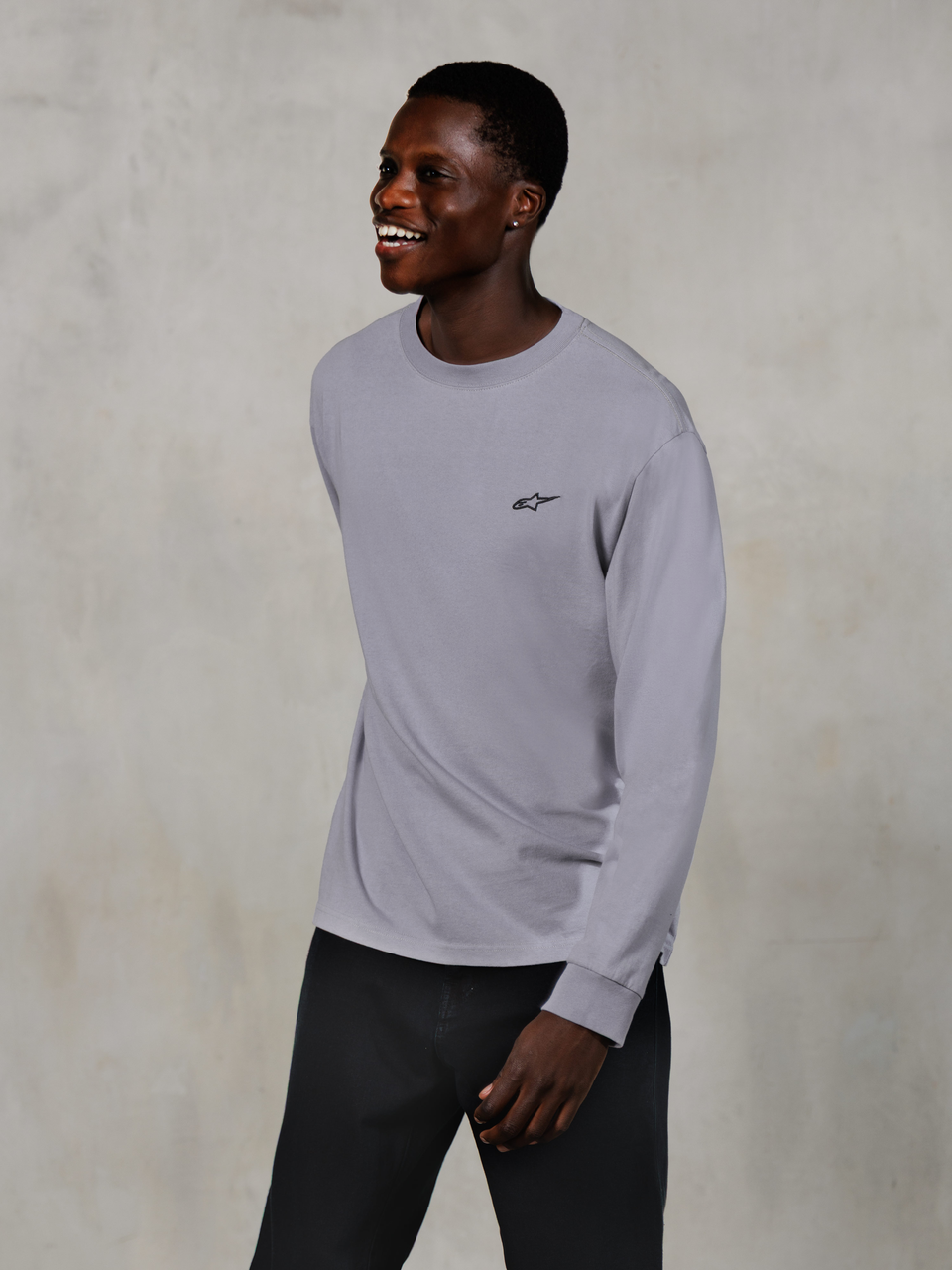 Alpinestars Essentials Ageless LS Tee, långärmad t-shirt, silver, liten svart logotyp på bröstet, bärs av en leende modell med mörka byxor, avslappnad modern passform, vardagliga motorsport-livsstilskläder