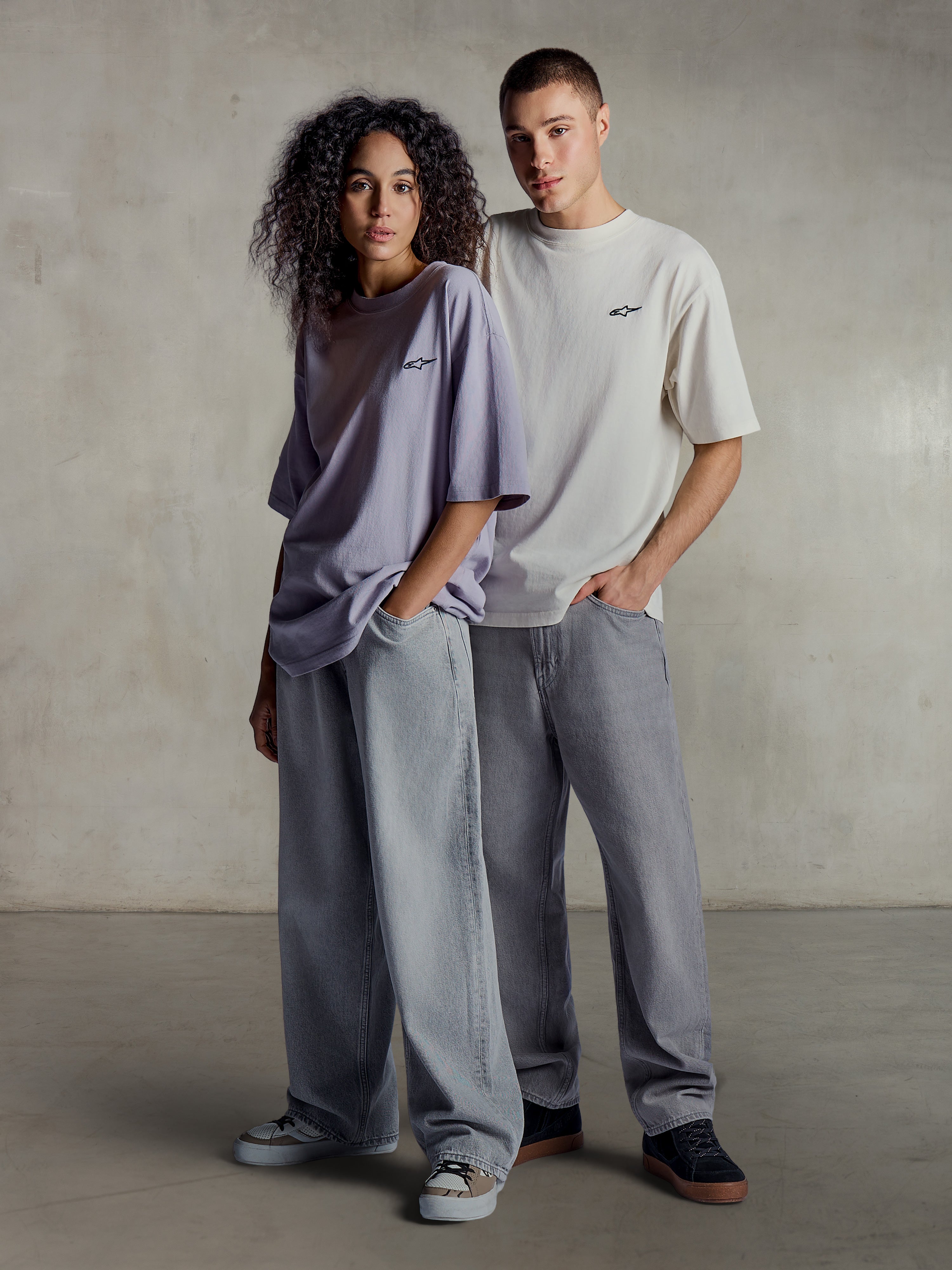 Alpinestars Essentials Ageless SS Tee, livsstilsbild på två modeller som bär kortärmade t-shirts i ljuslila och silver, avslappnad passform i ekologisk bomull med en subtil logotyp på bröstet, matchat med lösa jeans för en avslappnad streetwear-stil.