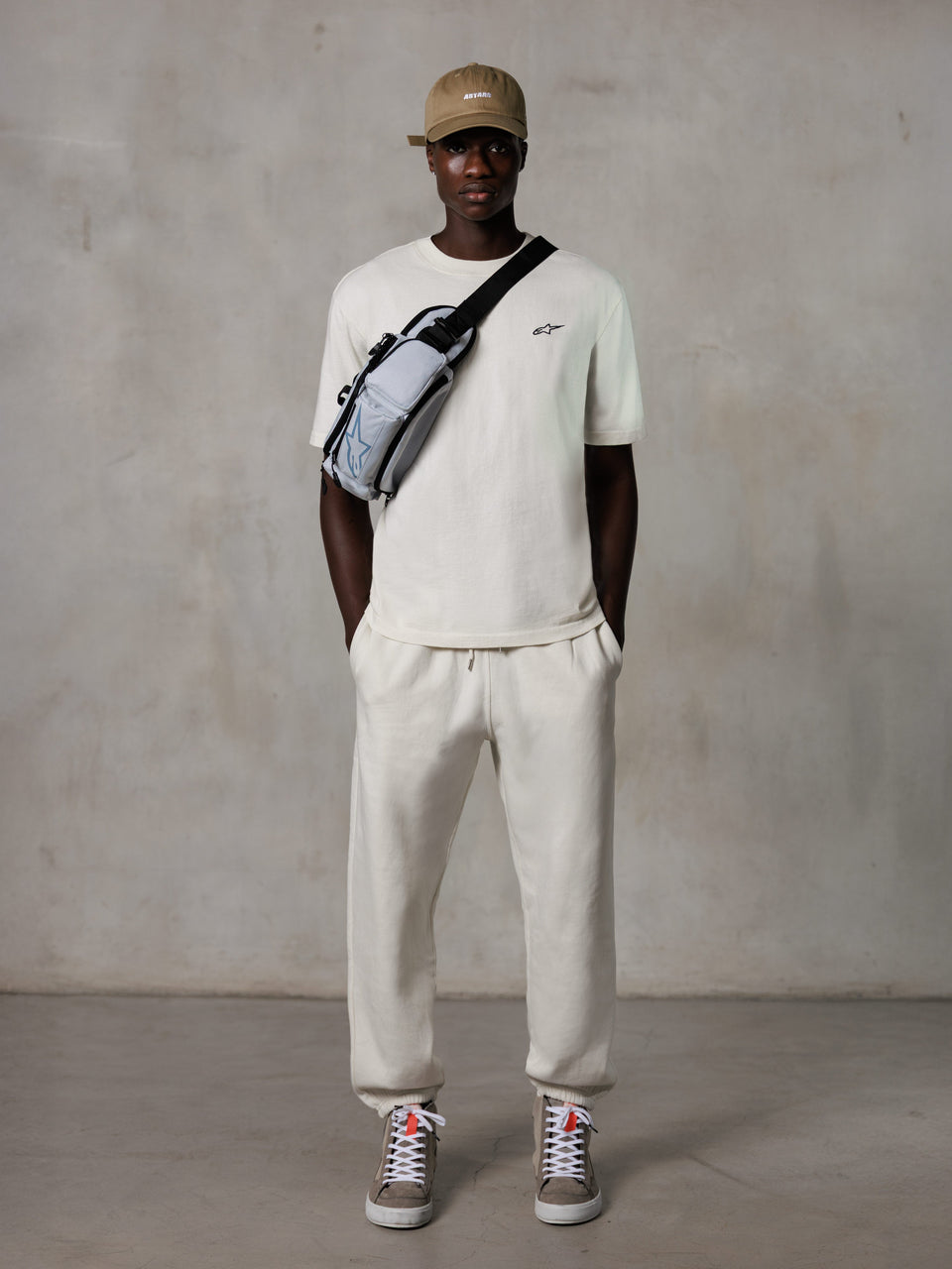 Alpinestars Essentials Ageless SS Tee, kortärmad t-shirt, Off White, med en liten svart logotyp på bröstet, bärs med matchande joggers, beige baseballkeps och en grå crossbody-väska, avslappnad lifestyle-streetwear