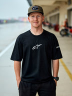 Man som bär Alpinestars Frontal CSF Tee, kortärmad T-shirt, svart, vit central Astars-logotyp, olivgrön Alpinestars-keps, avslappnat motorsportinspirerat streetwear