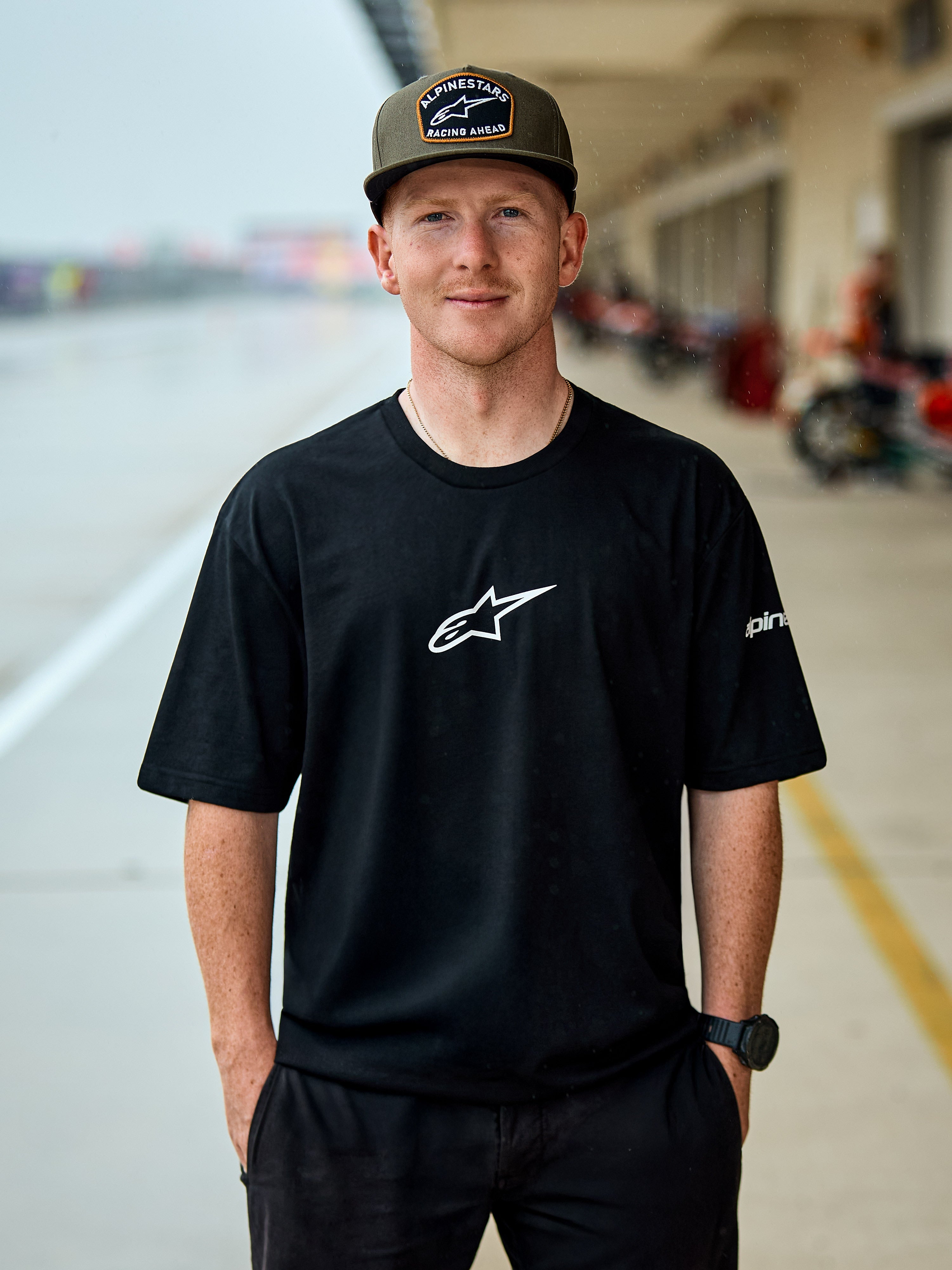 Man som bär Alpinestars Frontal CSF Tee, kortärmad T-shirt, svart, vit central Astars-logotyp, olivgrön Alpinestars-keps, avslappnat motorsportinspirerat streetwear