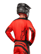 Motocrossförare sedd bakifrån bärande Alpinestars 2024 Fluid Lurv Jersey i Hot Orange och svart, med framträdande Alpinestars-logotyp på ryggen och ärmarna, kombinerad med matchande motocrossbyxor och en svart hjälm, designad för prestanda vid terrängkörning.