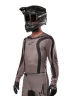 Alpinestars 2024 Fluid Lurv-tröja, motocrosströja, grå och svart, långärmad design med svängda svarta ränder och logotyper, bärs med matchande byxor och en svart motocrosshjälm, koordinerad racingutrustning för terrängkörning
