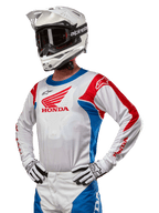 Motocrossförare i Honda Racer Iconic-tröja i vitt, klarblått och klarrött, med en stor Honda-vinglogotyp på bröstet, vit Alpinestars-hjälm och matchande vita och svarta handskar, koordinerad utrustning för offroad-prestanda