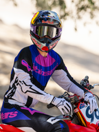 Motocrossförare på en röd motocrosscykel, Limited Edition HUNTA96 XXV Supertech Vision-glasögon i lila och vitt med silverspegel-linser, matchande lila, svart och vit Alpinestars-tröja och byxor, Red Bull-hjälm, vita handskar, koordinerad professionell motocrossutrustning